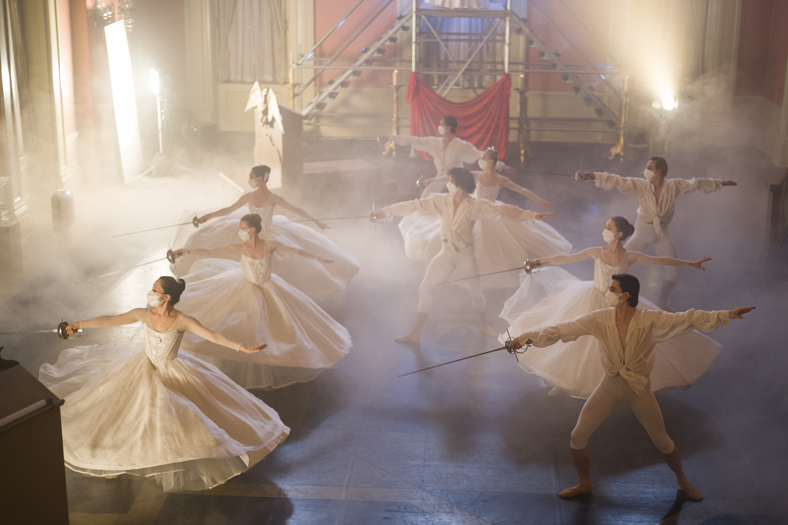 dancers on stage wear face masks and hold swords. Teh stage is foggy and they wear giant white dresses or white period clothing